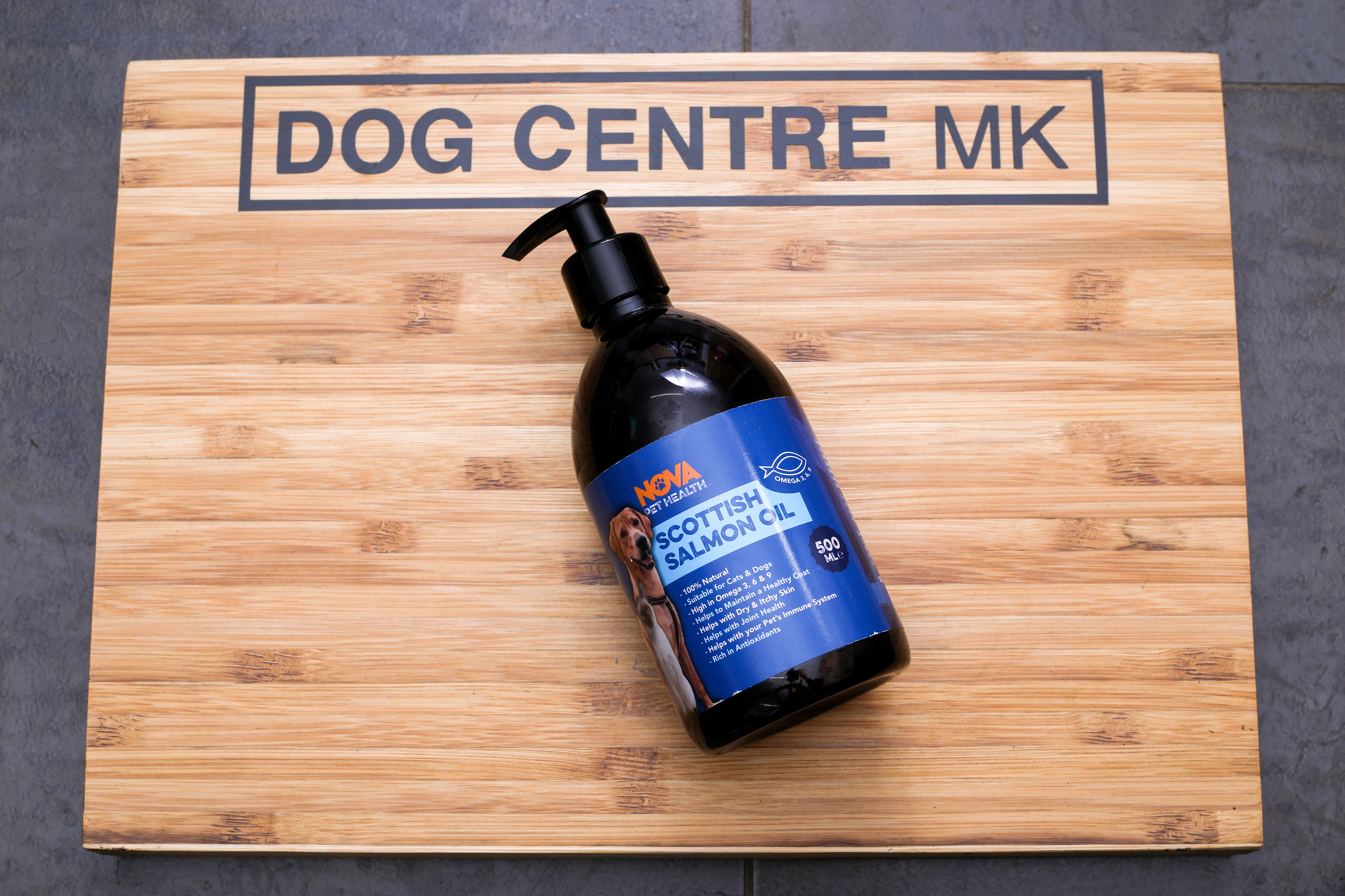 A 500ml bottle of Nova Scottish Salmon Oil for dogs, placed on a wooden surface with the brand name 'DOG CENTRE MK' visible in the background.