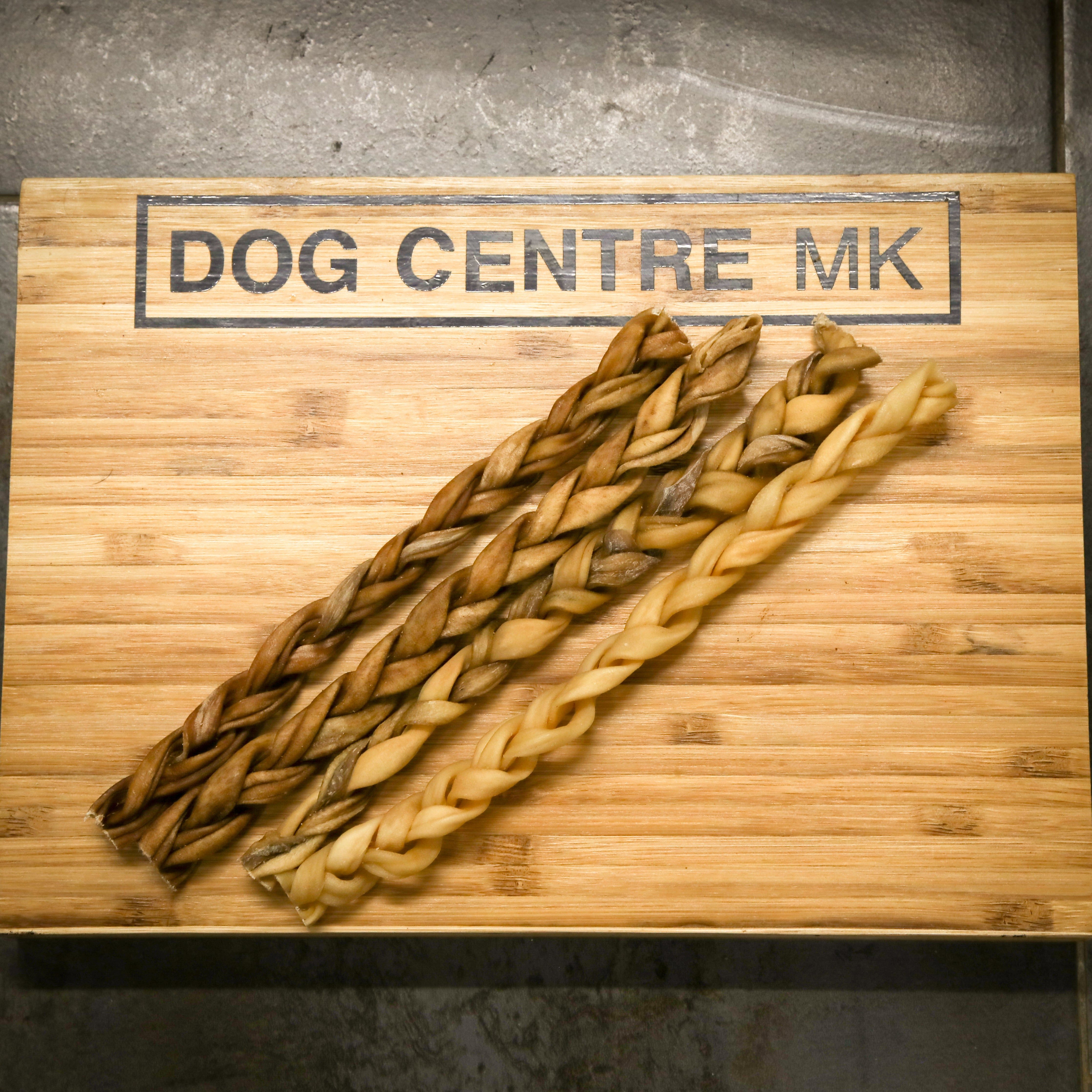 4 braided dog treats on a wooden board with 'DOG CENTRE MK' branding.