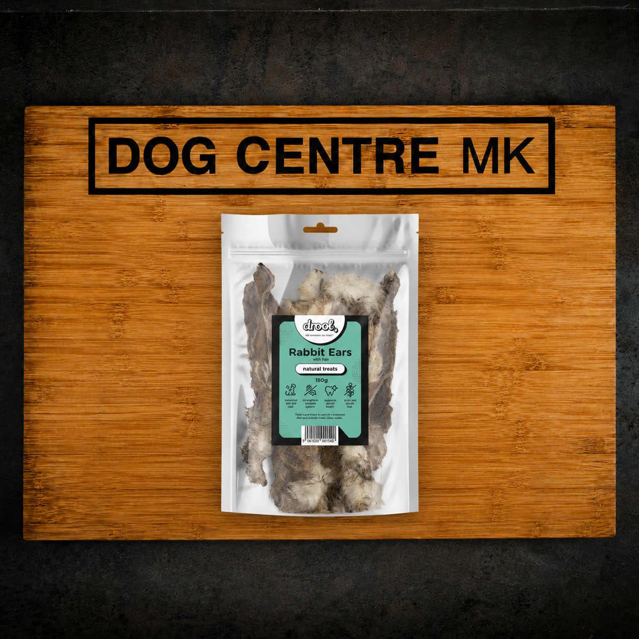 A 150g clear pack of Drool Natural Rabbit Ears with Hair dog treats, displayed on a wooden board branded with the Dog Centre MK logo.